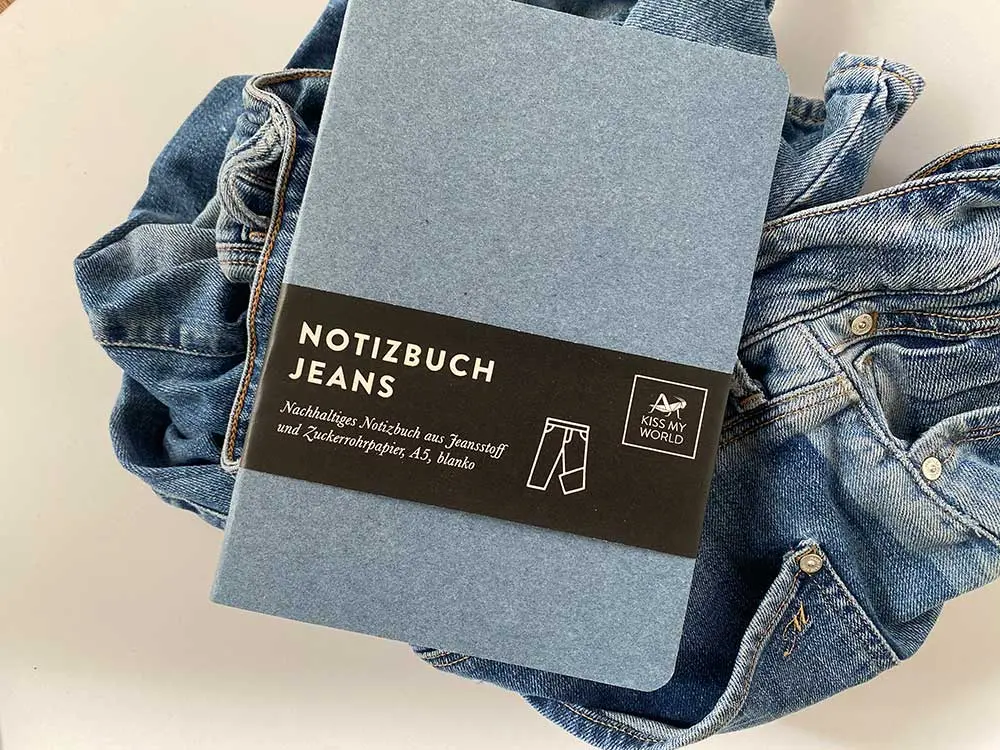 Jeans-Notizbuch A5, blanko mit Banderole, auf Jeansstoff, Produktfoto unseres Jeanspapier-Notizbuches auf einem Haufen mit Jeans-Stoffen und Jeanshosen für das Recycling. Dieser Stoff kann recycelt werden und daraus kann später Jeanspapier hergestellt werden und die Leute können unsere Papier à la Carte-Notizbücher kaufen