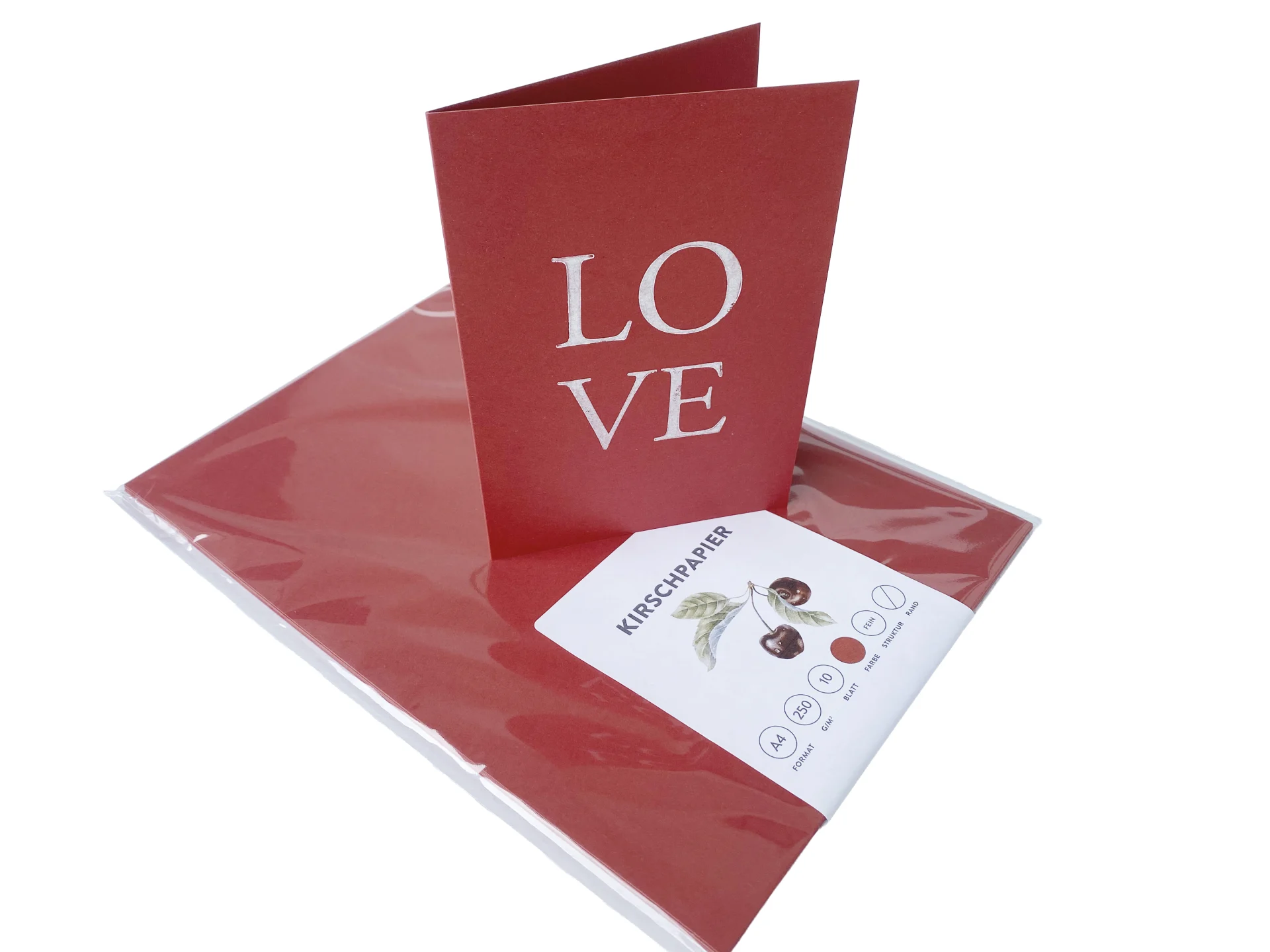 Ein in Klarsichtfolie verpacktes Set dunkelrotes Kirschpapier im A4-Format mit einer weißen Produktbanderole, auf dem eine aufgestellte, dunkelrote Klappkarte mit dem weißen Aufdruck LOVE steht.