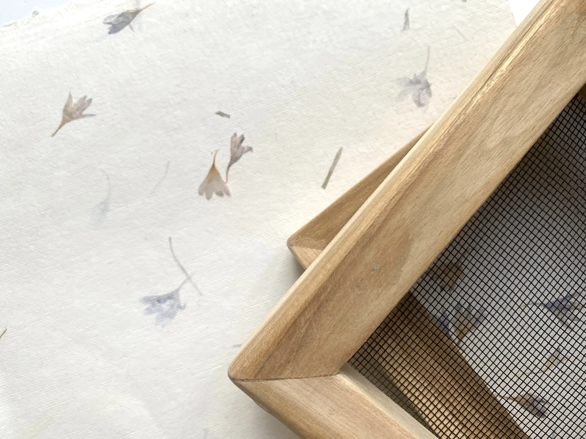 Holz-Schöpfrahmen mit Sieb neben handgeschöpftem Blütenpapier, helles Motiv mit Blüteneinschlüssen